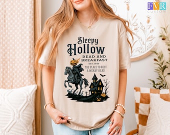 May include: Beige t-shirt featuring a Halloween-themed graphic. The design includes the text "Sleepy Hollow Dead and Breakfast" with a headless horseman, a haunted house, and pumpkins. The shirt is a relaxed fit.