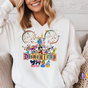 Puede incluir: Sudadera con capucha blanca con un colorido gr&aacute;fico de Disney Trip 2026. El dise&ntilde;o incluye a Mickey Mouse, Minnie Mouse, el Pato Donald y otros personajes de Disney, junto con un castillo y fuegos artificiales.