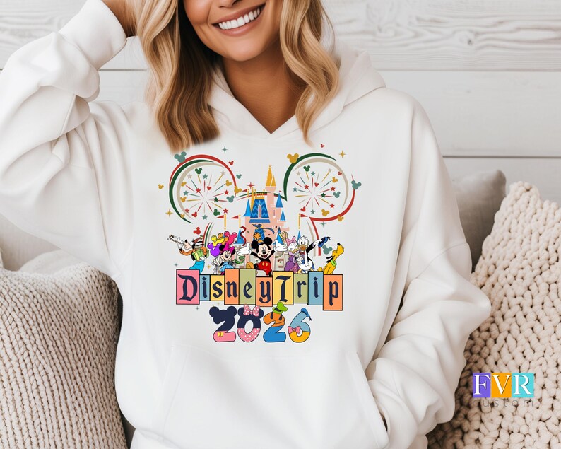 Puede incluir: Sudadera con capucha blanca con un colorido gr&aacute;fico de Disney Trip 2026. El dise&ntilde;o incluye a Mickey Mouse, Minnie Mouse, el Pato Donald y otros personajes de Disney, junto con un castillo y fuegos artificiales.