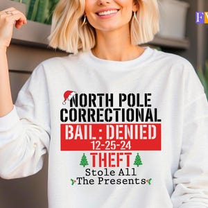May include: A white long-sleeve sweatshirt featuring black and red text that reads NORTH POLE CORRECTIONAL BAIL: DENIED 12-25-24 THEFT Stole All The Presents. The design includes a small red Santa hat, two green Christmas trees, and holly icons.