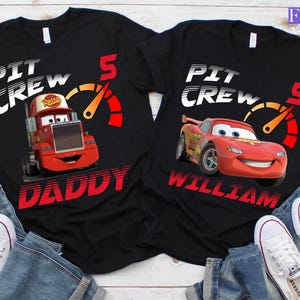 May include: Black t-shirts featuring the text "PIT CREW" and a speedometer graphic. One shirt has a red cartoon truck with "DADDY" below, the other has a red race car with "WILLIAM" below. Both have the number 5.