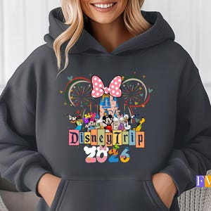 Puede incluir: Sudadera con capucha gris oscuro con un dise&ntilde;o colorido de Disney Trip 2023. El dise&ntilde;o incluye un lazo rosa con lunares, un castillo, fuegos artificiales y varios personajes de Disney. La sudadera tiene una capucha con cord&oacute;n y un bolsillo delantero.