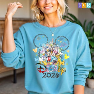 May include: A turquoise sweatshirt with a Disney design. The design features Mickey Mouse, Minnie Mouse, Donald Duck, Daisy Duck, Goofy, and Pluto, a castle, fireworks, and the text "Disney 2026".