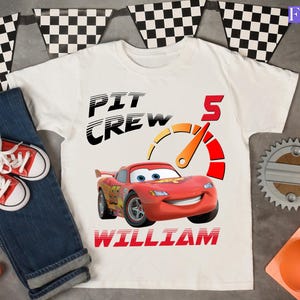 May include: White t-shirt with a graphic of a red race car, the text "PIT CREW", the number "5", and the name "WILLIAM". The shirt is paired with blue jeans and red sneakers. Checkered flags are in the background.