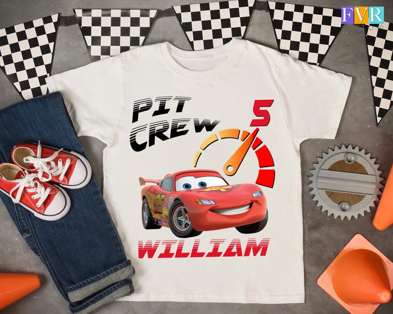 May include: White t-shirt with a graphic of a red race car, the text "PIT CREW", the number "5", and the name "WILLIAM". The shirt is paired with blue jeans and red sneakers. Checkered flags are in the background.