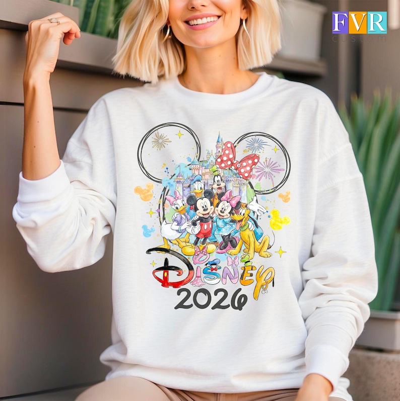 May include: White long-sleeved sweatshirt with a vibrant Disney-themed graphic. The design showcases Mickey and Minnie Mouse, Donald and Daisy Duck, and Goofy, alongside a castle, fireworks, and the text "Disney 2026".