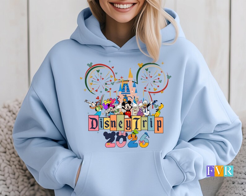 Disney Trip 2026 hoodie with Mickey Mouse, Minnie Mouse and Disney friends in front of a colorful castle with fireworks inside Minnie ears silhouette on blue pullover hoodie.