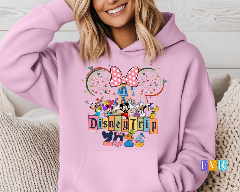 Puede incluir: Sudadera con capucha rosa claro con un dise&ntilde;o de Disney Trip 2023. El dise&ntilde;o incluye a Mickey y Minnie Mouse, otros personajes de Disney, un lazo de lunares y una ilustraci&oacute;n de castillo.