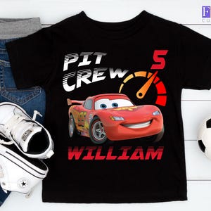 May include: Black t-shirt featuring a graphic of Lightning McQueen from the movie Cars, with the text "PIT CREW" and "5". The name "WILLIAM" is printed below the car. The image includes jeans, white sneakers, and a soccer ball.