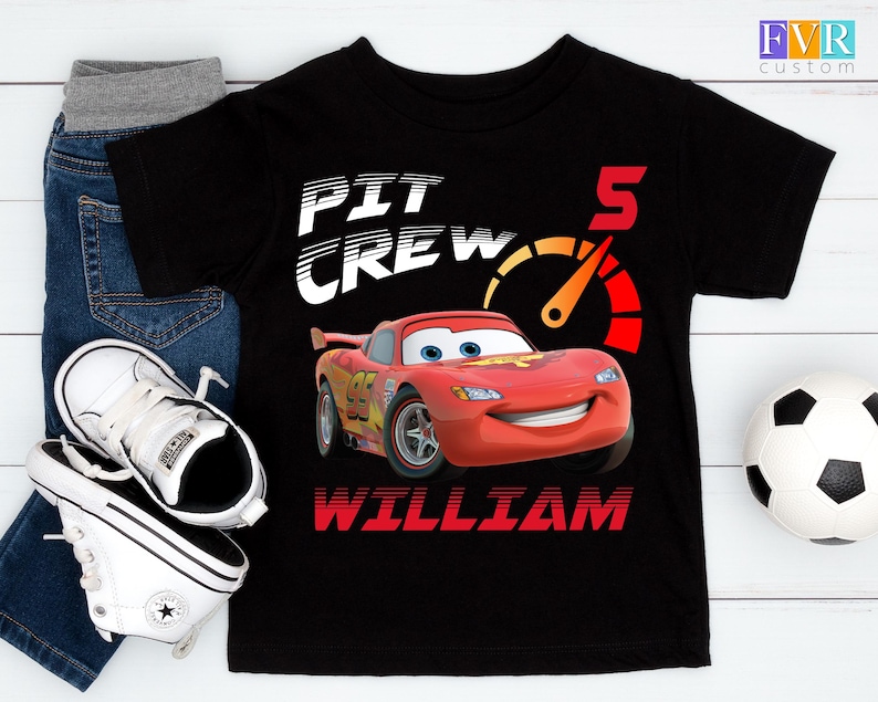 May include: Black t-shirt featuring a graphic of Lightning McQueen from the movie Cars, with the text "PIT CREW" and "5". The name "WILLIAM" is printed below the car. The image includes jeans, white sneakers, and a soccer ball.