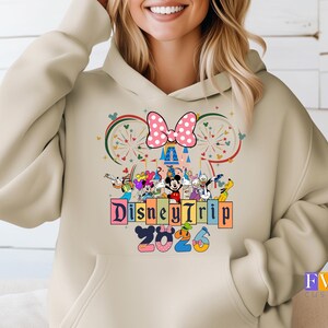 Puede incluir: Sudadera con capucha beige con un dise&ntilde;o de Disney Trip 2023. El dise&ntilde;o incluye a Mickey y Minnie Mouse, otros personajes de Disney, un lazo rosa con lunares y un castillo. La sudadera tiene un bolsillo delantero y una capucha con cord&oacute;n.