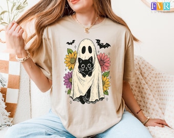 May include: Beige t-shirt featuring a Halloween-themed graphic. The design includes a ghost holding a black cat, surrounded by colorful flowers and bats. The graphic is centered on the shirt, creating a whimsical and festive look. The shirt is made of a soft material.