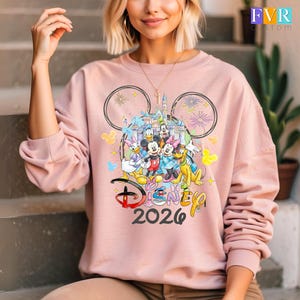 May include: A pale pink sweatshirt with a Disney design. The design includes Mickey Mouse, Minnie Mouse, Donald Duck, Daisy Duck, Goofy, and Pluto, with a castle backdrop. The text "Disney 2026" is also visible.