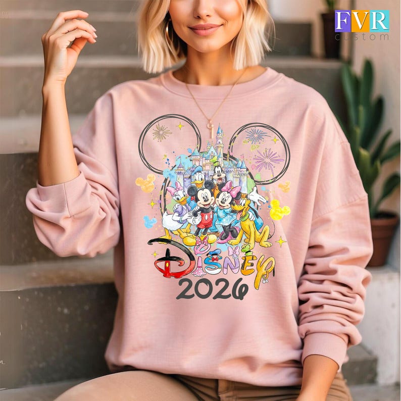 May include: A pale pink sweatshirt with a Disney design. The design includes Mickey Mouse, Minnie Mouse, Donald Duck, Daisy Duck, Goofy, and Pluto, with a castle backdrop. The text "Disney 2026" is also visible.