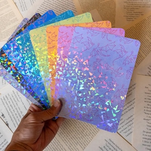 Puede incluir: Una colección de tarjetas rectangulares con un patrón repetitivo de murciélagos. Cada tarjeta muestra un degradado de color distinto, incluyendo morado, naranja, amarillo, verde y azul, con un efecto holográfico de cristal roto.