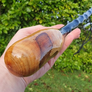 May include: A handcrafted wooden walking stick with a polished, light brown body and a dark blue, twisted handle. The wood grain is visible, and a black cord is attached. The stick is held in a hand against a green background.