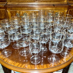 May include: A collection of clear glass stemware with gold rims, including wine glasses, champagne flutes, and other drinkware. The glasses are arranged on a round, wooden table, showcasing a vintage aesthetic.