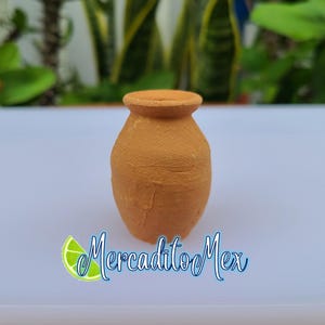 May include: A small, handcrafted terracotta vase with a rounded body and a flared rim. The vase is a warm, earthy brown color and sits on a white surface. The background features green foliage.