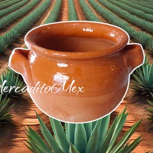 May include: A brown ceramic pot with two handles, set against a backdrop of agave plants. The pot has a glossy finish and a wide opening. The text "Mercadito Mex" is written in white script on the pot.