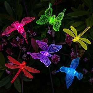 May include: A collection of colorful, illuminated garden stakes featuring various insect and bird designs. The stakes include a pink hummingbird, a green butterfly, a yellow dragonfly, a red dragonfly, a purple butterfly, and a blue hummingbird. The stakes are set against a backdrop of green foliage and small pink flowers.