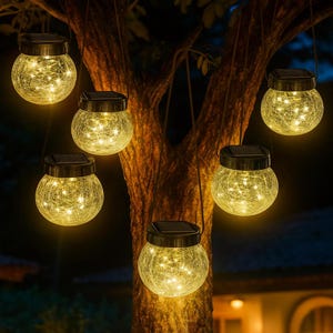 May include: Several solar-powered, crackle-glass jar lights hang from a tree. Each jar has a black lid with a solar panel and contains warm white LED lights. The lights are suspended by black cords, creating a decorative outdoor lighting display.
