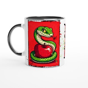 May include: White ceramic mug with a cartoon illustration of a green snake coiled around a red apple on a red background. The mug has a black rim and handle, with black accents and splatters.
