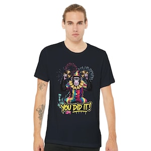 Op de afbeelding: Zwart t-shirt met een kleurrijke illustratie van een aap verkleed als clown, die bekkens vasthoudt. De aap draagt een regenboogoutfit en een gerimpelde kraag. De tekst "YOU DID IT!" staat eronder.