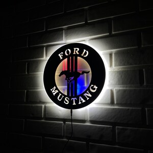 May include: A black metal wall sign with a white outline and the words "FORD MUSTANG" in white lettering. The sign features a red and blue illuminated horse and bar logo.