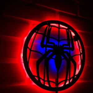 Op de afbeelding: Een zwart silhouet van een spinnenweb met een spin in het midden, verlicht door rode en blauwe LED-verlichting. De lamp is gemonteerd op een bakstenen muur.