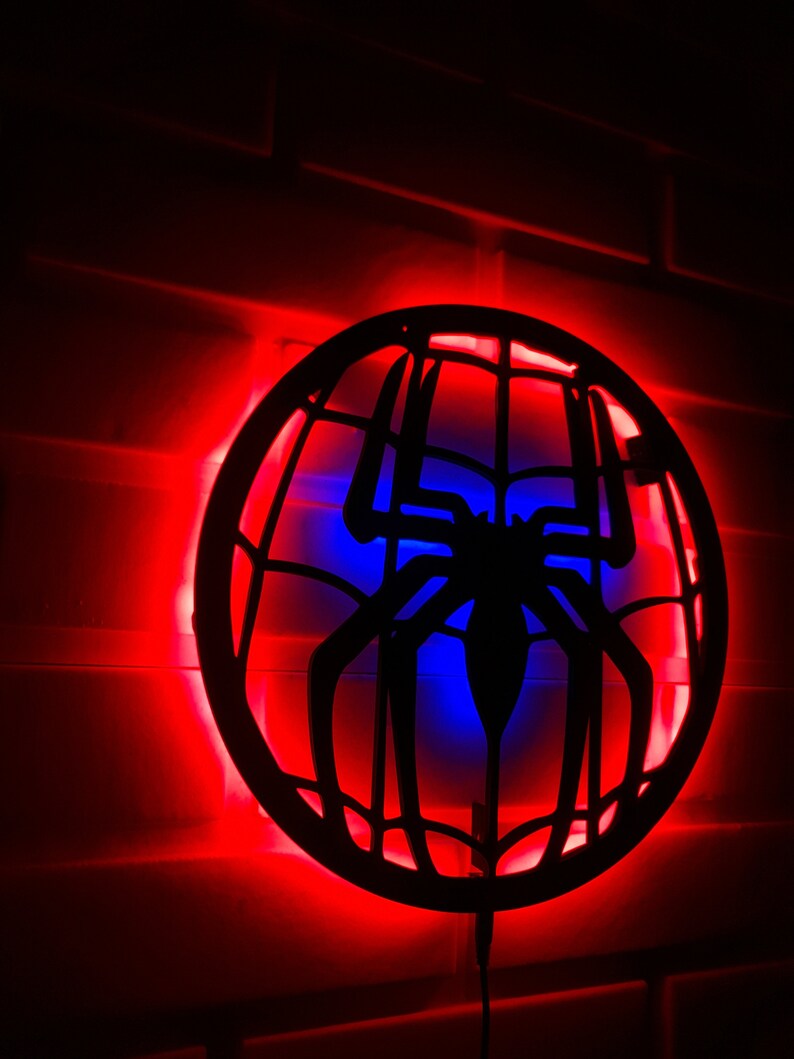 Op de afbeelding: Een zwart silhouet van een spinnenweb met een spin in het midden, verlicht door rode en blauwe LED-verlichting. De lamp is gemonteerd op een bakstenen muur.