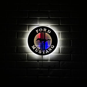 May include: A black and white Ford Mustang logo with red, white, and blue accents. The logo is illuminated and mounted on a white brick wall.