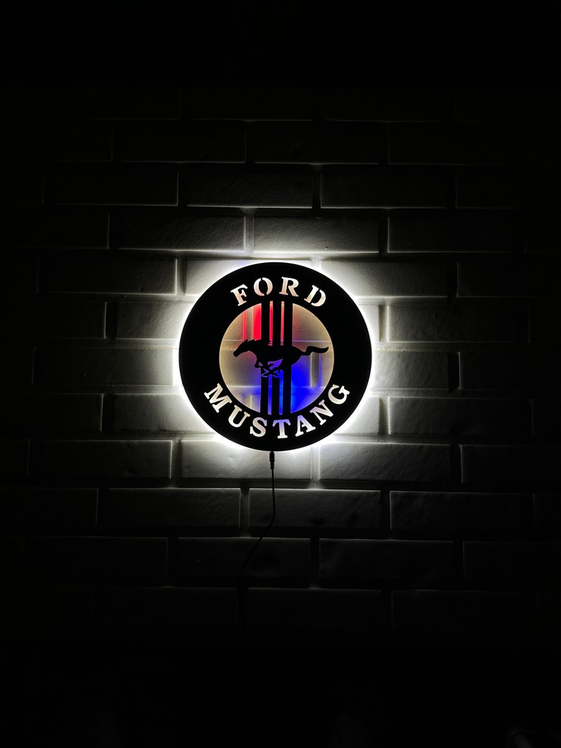 May include: A black and white Ford Mustang logo with red, white, and blue accents. The logo is illuminated and mounted on a white brick wall.