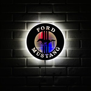May include: A black and white Ford Mustang logo with red, white, and blue stripes. The logo is illuminated with a white light and is mounted on a white brick wall.
