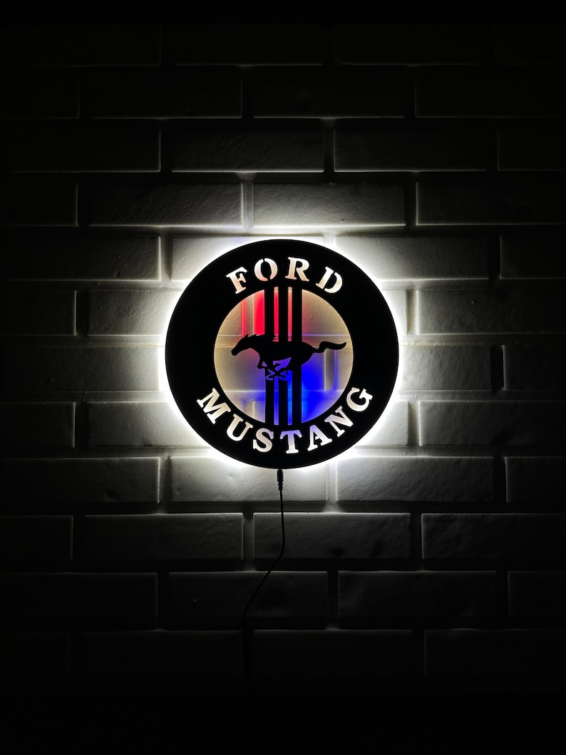 May include: A black and white Ford Mustang logo with red, white, and blue stripes. The logo is illuminated with a white light and is mounted on a white brick wall.