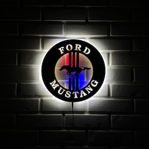 May include: A black metal sign with a white outline and a red, white, and blue LED light behind it. The sign features the Ford Mustang logo with the words "FORD" and "MUSTANG" in white lettering.