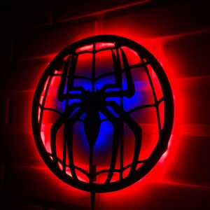 Op de afbeelding: Een zwart silhouet van een spinnenweb met een spin in het midden, verlicht door rode en blauwe LED-verlichting. De lamp is gemonteerd op een witte bakstenen muur.