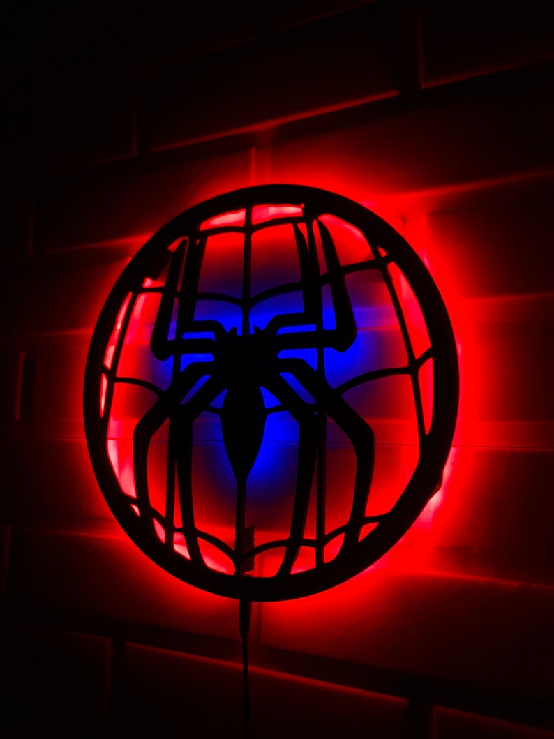 Op de afbeelding: Een zwart silhouet van een spinnenweb met een spin in het midden, verlicht door rode en blauwe LED-verlichting. De lamp is gemonteerd op een witte bakstenen muur.
