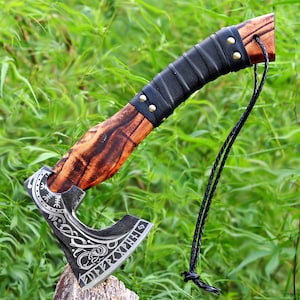 Könnte beinhalten: Eine handgefertigte Axt mit einem Holzgriff mit dunklem Maserungsmuster. Der Griff ist mit schwarzem Leder umwickelt und mit Messingnieten befestigt. Der Axtkopf ist silberfarben mit geätzten Designs und einer scharfen Klinge. Eine schwarze Schnur ist befestigt.