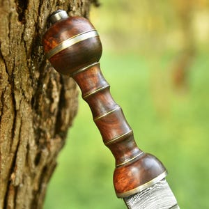 May include: A sword handle featuring a polished dark brown wooden grip with a rounded pommel and guard. The handle is accented with thin metallic rings separating the grip sections, and it connects to a patterned Damascus steel blade.