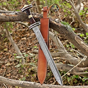 May include: A double-edged sword with a patterned Damascus steel blade, a dark wood handle with light-coloured bone or horn inlays, and a tan leather scabbard featuring a belt loop and a metal snap closure, resting against a fallen tree branch.