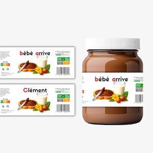 Peut inclure: Un pot de pâte à tartiner au chocolat et aux noisettes avec un couvercle blanc. L'étiquette indique "bébé arrive" avec une illustration de pain grillé, un verre de lait et des fruits. Deux étiquettes supplémentaires sont également présentes.