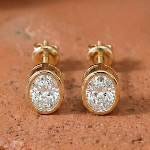 Könnte beinhalten: Ein Paar goldene Ohrstecker mit ovalen, klaren Edelsteinen. Die Ohrringe sind in einer Lünettenfassung gefasst und werden auf einer strukturierten, rotbraunen Oberfläche präsentiert. Klassischer Schmuck.