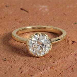 Puede incluir: Un anillo de oro con una gran piedra central de diamante transparente y redondo. El diamante está engastado en un engaste de bisel, y la banda es lisa y pulida. El anillo está sobre una superficie marrón rojiza.