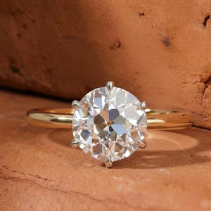 May include: A gold ring with a large, round, clear diamond. The diamond is held in place by six prongs. The ring is set against a reddish-brown background.