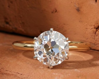 May include: A gold ring with a large, round, clear diamond. The diamond is held in place by six prongs. The ring is set against a reddish-brown background.