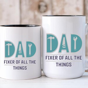 May include: Two white ceramic mugs with black handles and rims. The word "DAD" is printed in teal with tool graphics. Below, the text reads "FIXER OF ALL THE THINGS" in a dark blue font. The mugs are of different sizes.