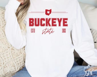 May include: White long-sleeved sweatshirt featuring the word "BUCKEYE" in red block letters. Above the word is a red Ohio state outline with a buckeye leaf. Below the word are the words "ESTO state 1803" in red script.