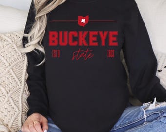 May include: Black long-sleeve sweatshirt featuring the red text "BUCKEYE state". The design includes the Ohio state outline and the year 1803. A casual apparel item.