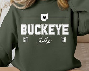 May include: Dark green sweatshirt featuring the word "BUCKEYE" in white capital letters. Above the text is a white outline of the state of Ohio with a buckeye leaf. Below the text are the words "ESTD state 1803" in white.