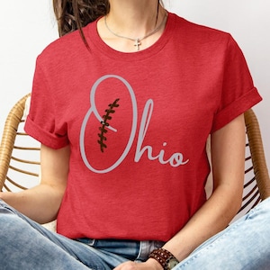 May include: Red t-shirt with the word "Ohio" in a script font, with a football graphic. The shirt has rolled-up sleeves and is made of a soft material. The design is in gray.